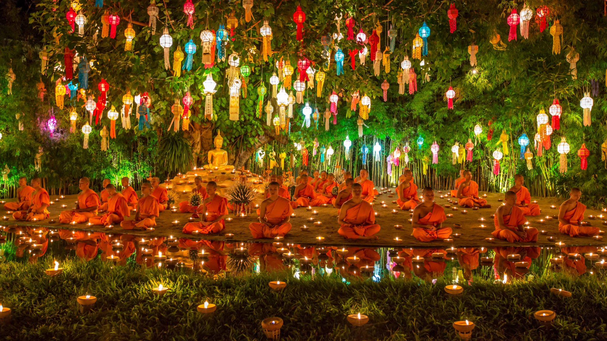 Dużo mnichów ubranych w pomarańczowe szaty siedzących nad wodą z zapalonymi świecami modlących się, na drzewach dużo kolorowych latarni, figura Buddy pod drzewem