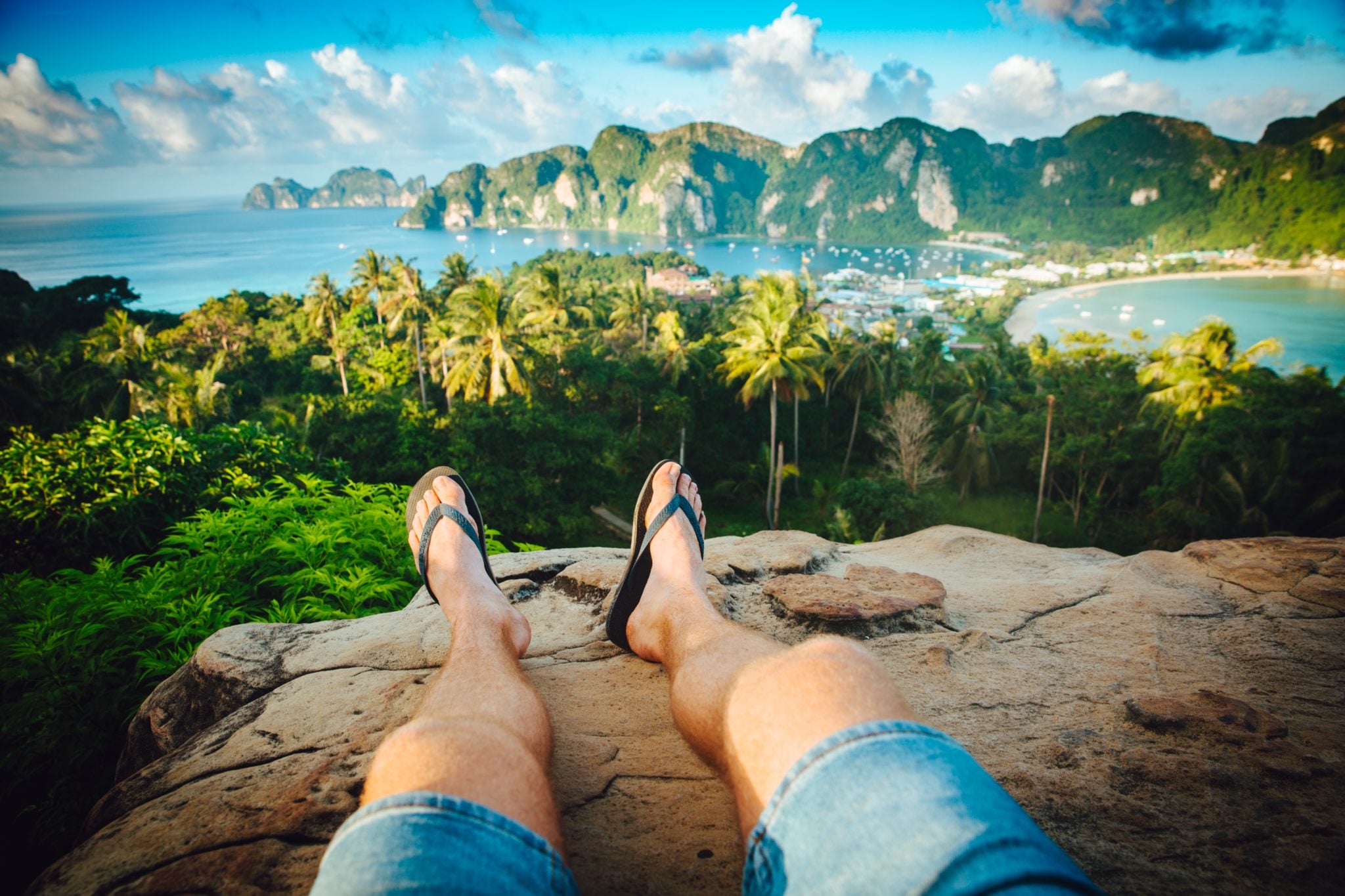 Mężczyzna siedzący na skale na punkcie widokowym w Tajlandii w słoneczny dzień, z widokiem na morze, łodzie, klify, wyspy i palmy