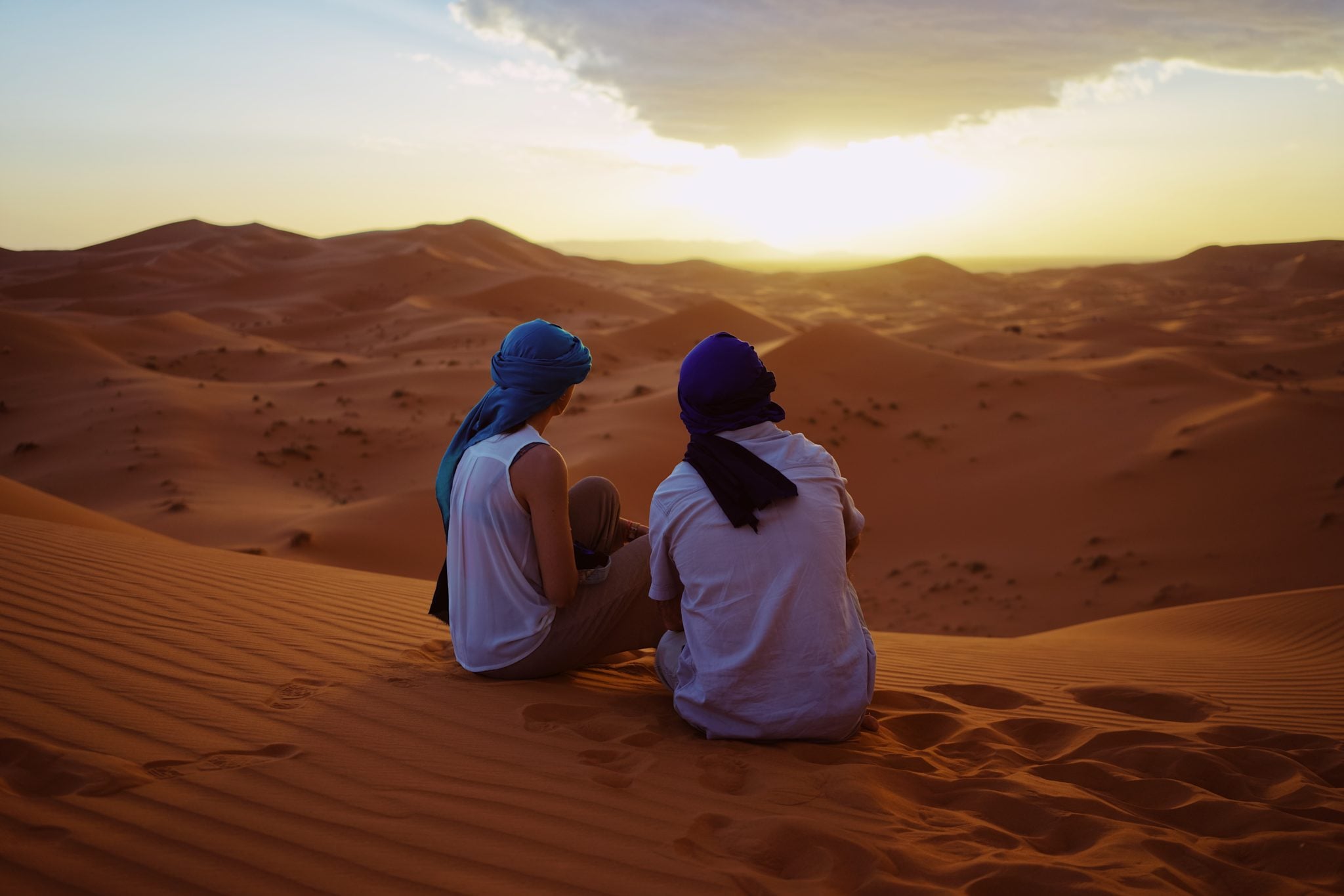 Chłopak i dziewczyna z niebieskimi turbanami na głowie, siedzący na wydmie na pustyni o zachodzie słońca