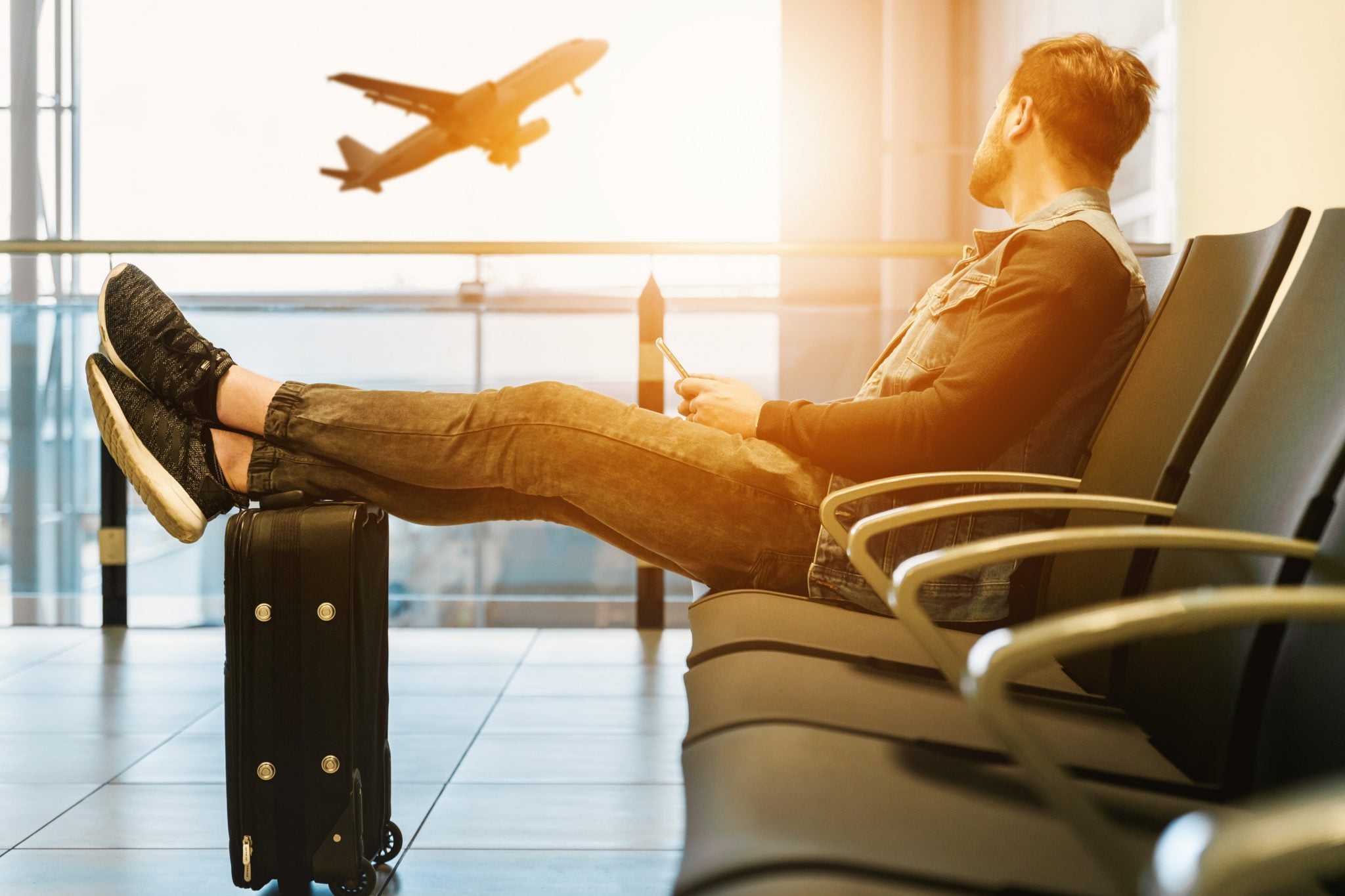 Chłopak na lotnisku w hali odlotów, siedzący i czekający na swój lot, trzyma nogi na walizce i obserwuje startujący samolot o zachodzie słońca