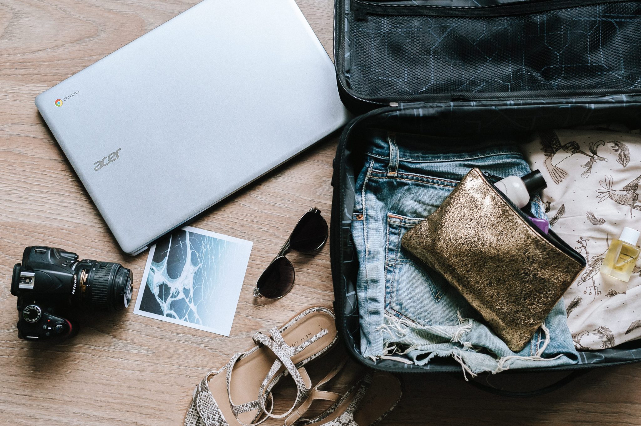 Rozpięta walizka leżąca na ziemi, obok ubrania, okulary przeciwsłoneczne i laptop