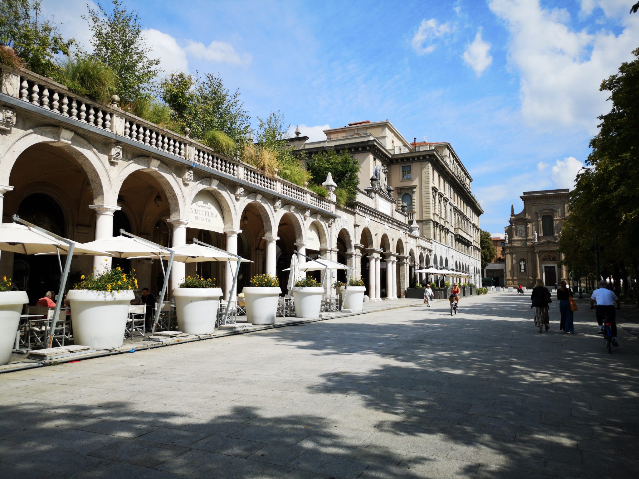 Budynek arkadowy w miasteczku Bergamo we Włoszech w słoneczny dzień, na zewnątrz stoliki kawiarni i restauracji