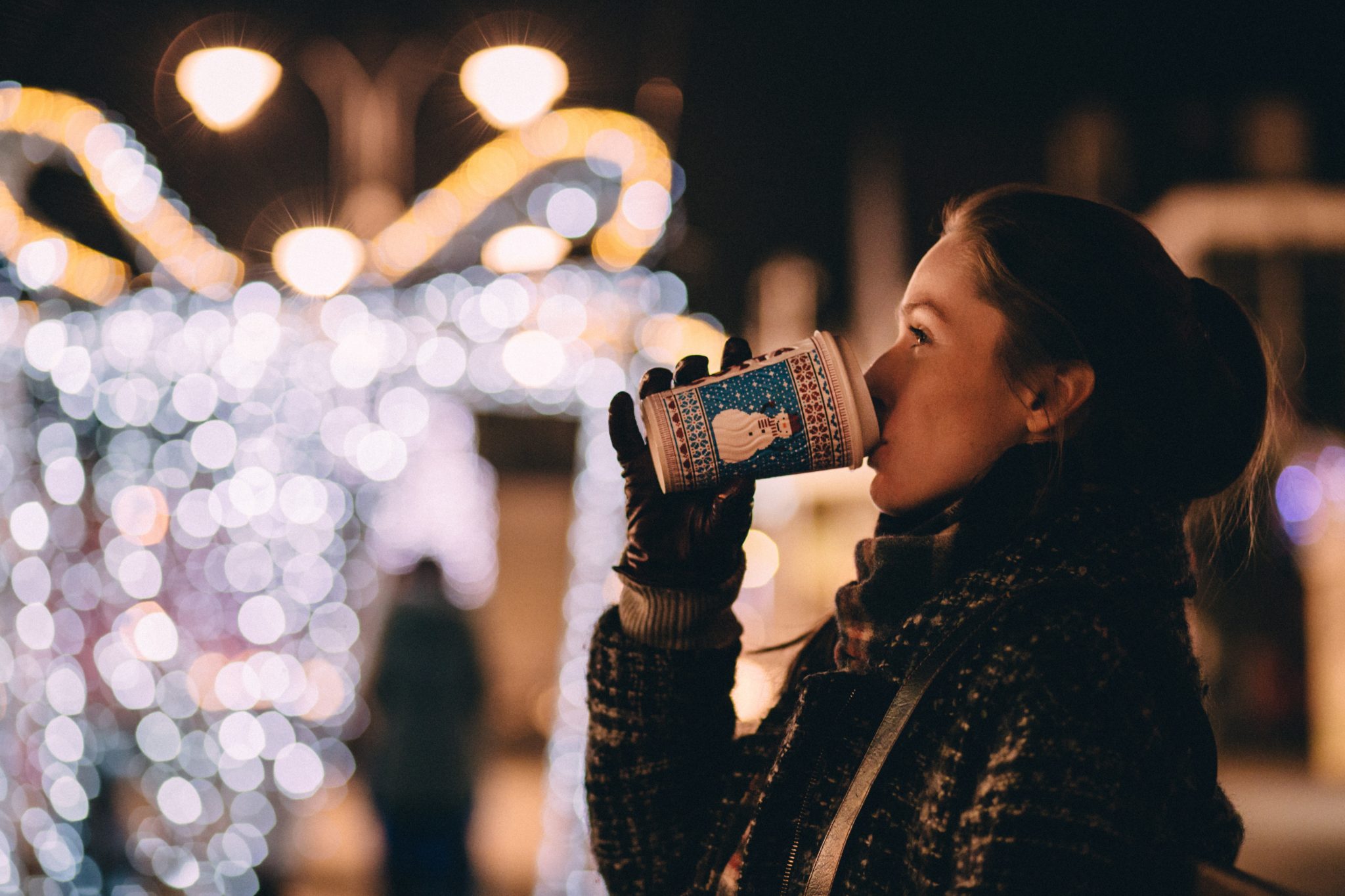 Dziewczyna ciepło ubrana w rękawiczkach i szaliku, pijąca grzane wino z kubeczka na markecie świątecznym w zimowej scenerii