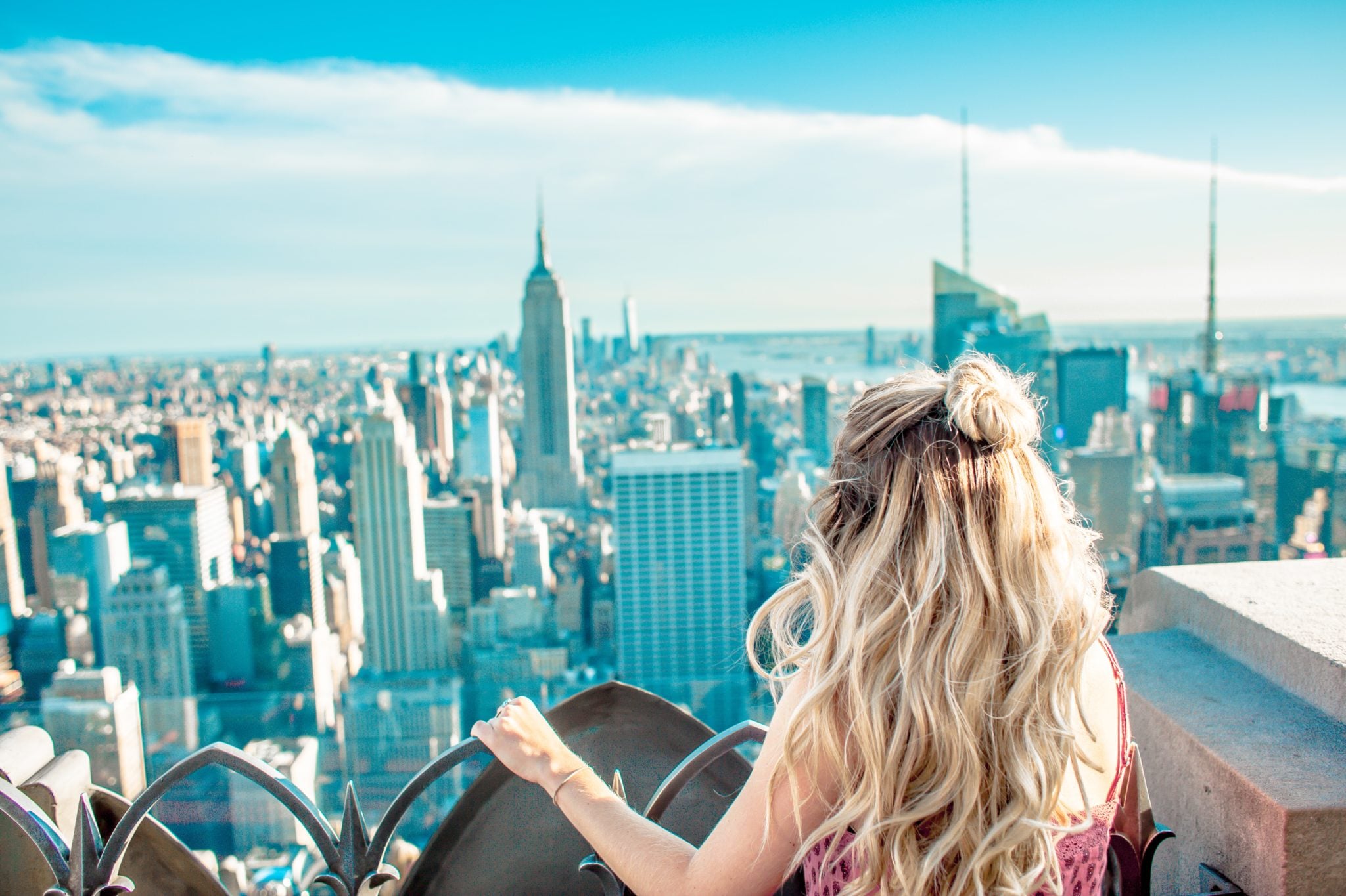 Dziewczyna stojąca na tarasie widokowym obserwująca panoramę Nowego Jorku. Bardzo dużo budynków, wieżowców, niebieskie niebo, słoneczny dzień.