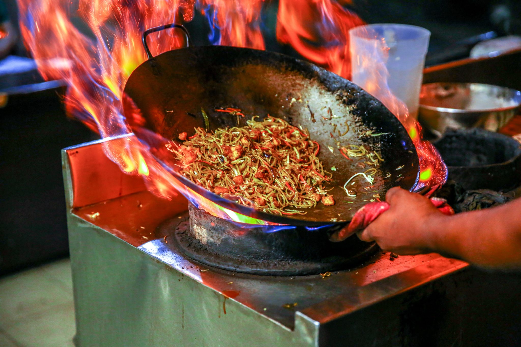 Mężczyzna smażący noodle metodą stir fry w dużym woku wypełnionym noodlami i warzywami na dużym ogniu