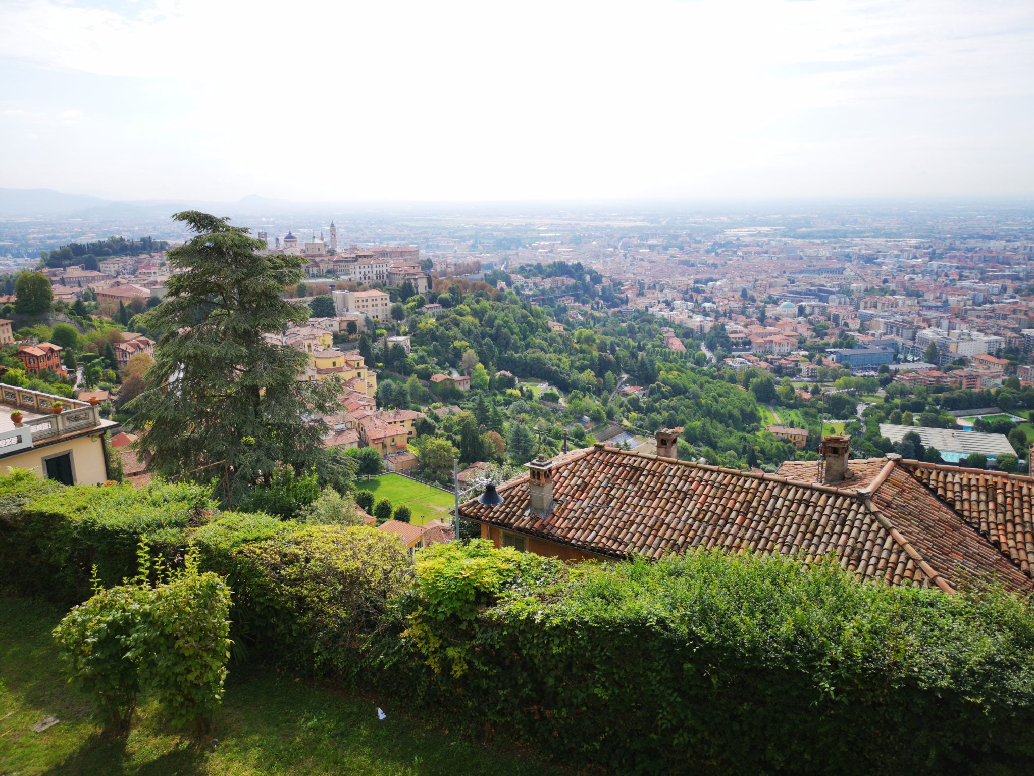 Panoramiczny widok na stare miasto w Bergamo, na kolorowe domki, stare budynki i dużo zieleni, w tle Alpy i budynki Mediolanu