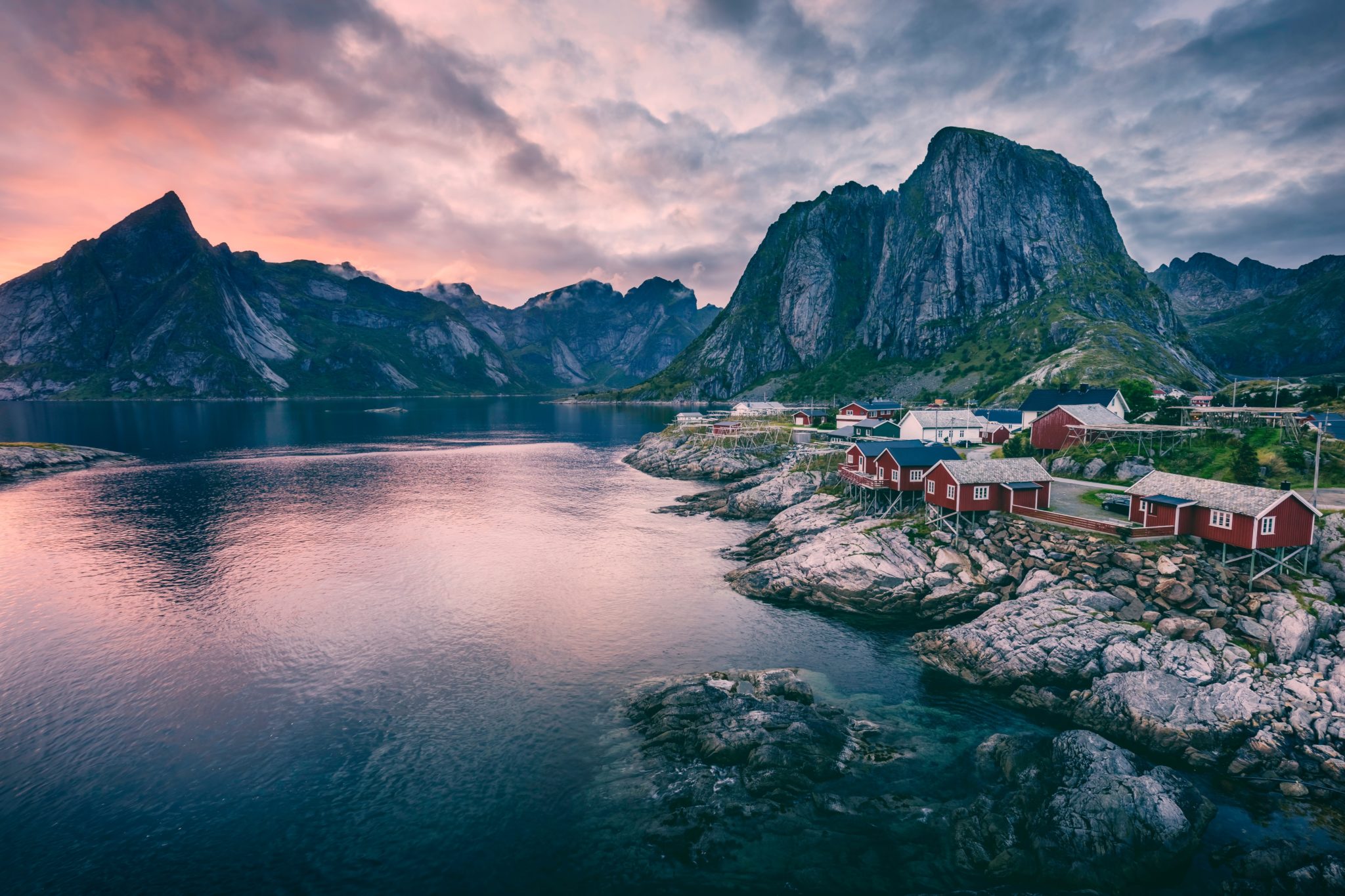Widok na małą, rybacką wioskę w Norwegii otoczoną fiordami o zachodzie słońca. Kolorowe rybackie domki odbijają się w tafli wody.