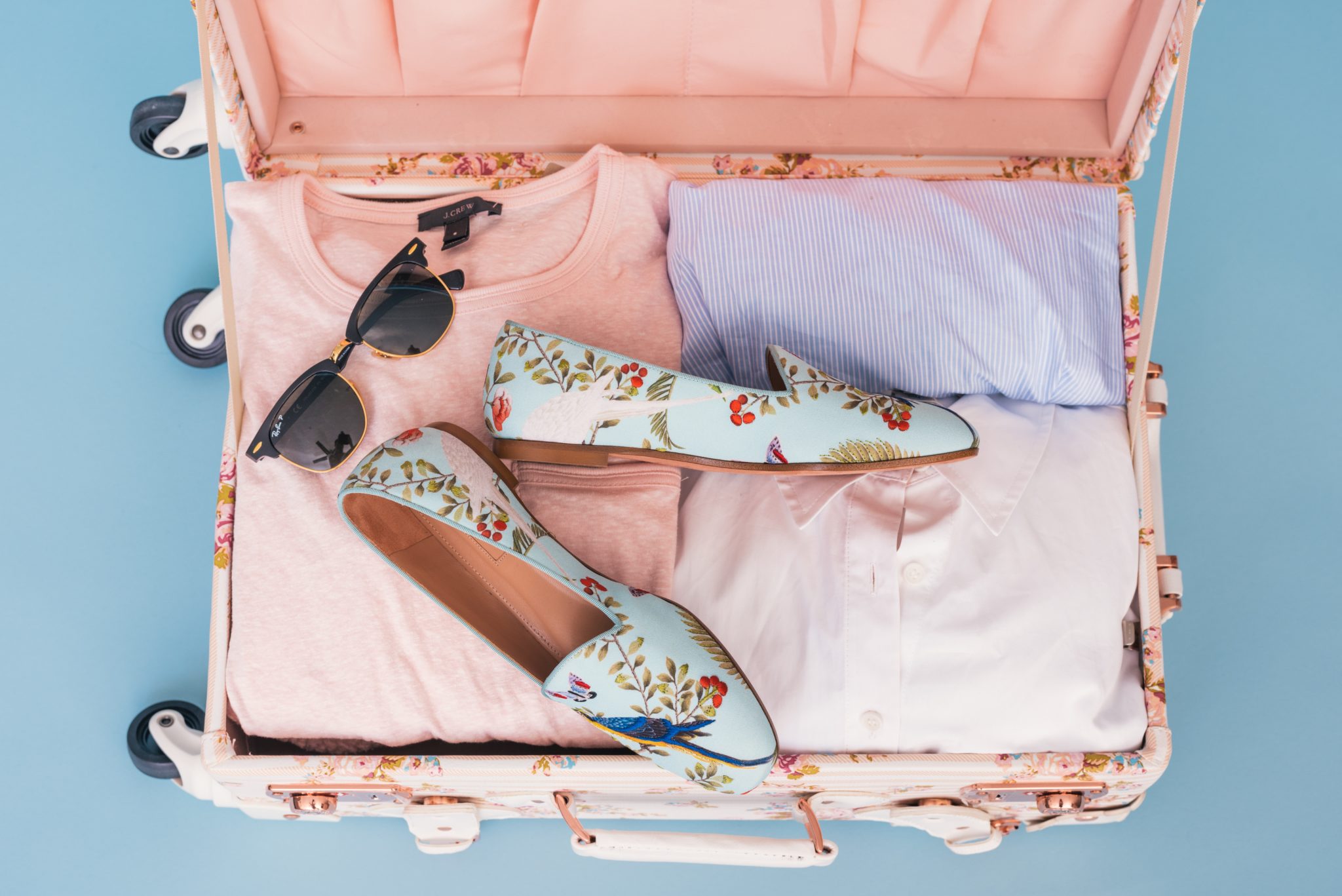 Otwarta różowa walizka, w trakcie pakowania, okulary przeciwsłoneczne, kolorowe buty w kwiaty, różowa koszulka, niebieska koszulka